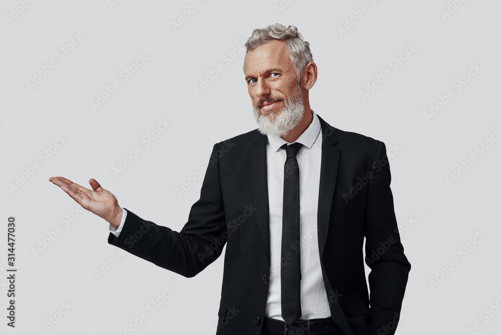 Confident mature man in full suit pointing copy space and looking at camera while standing against grey background Confident mature man in full suit pointing copy space and looking at camera while standing against grey background