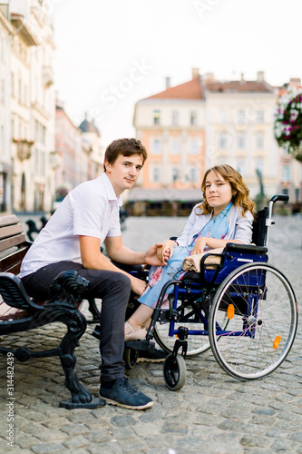 Young man and woman in wheelchair have fun and smiling on the bench outside. Old city street, love and disability concept