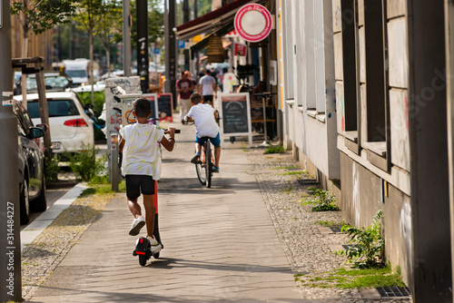Canvas Print A little boy drives an e-scooter, in the background a boy with a bike, sunny day