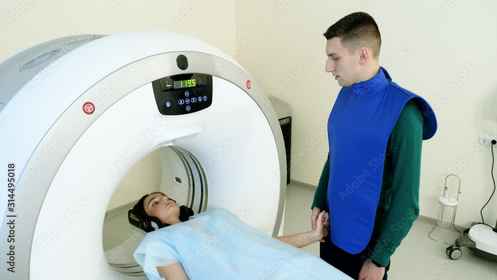 Procedure of computed tomography or magnetic resonance imaging. Female patient on medical examination. CT or MRI scan. Handsome man holding hand of his girlfriend to comfort her. 4K Procedure of computed tomography or magnetic resonance imaging. Female patient on medical examination. CT or MRI scan. Handsome man holding hand of his girlfriend to comfort her. 4K