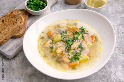 Fish soup made of salmon and vegetables in white bowl, served with bread toasts, parsley, lemon on textured grey table. Vegetarian and healthy food rich with Omega 3.