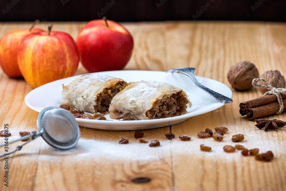 Fototapeta premium Apple roll pie - Apple Strudel. Pie with apples, cinnamon, walnuts and raisins, sprinkled with powdered sugar in a plate on a wooden table 