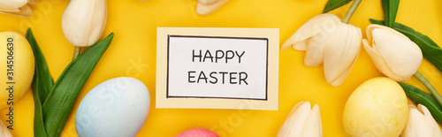 top view of tulips and painted Easter eggs around card with happy Easter lettering on colorful yellow background, panoramic shot