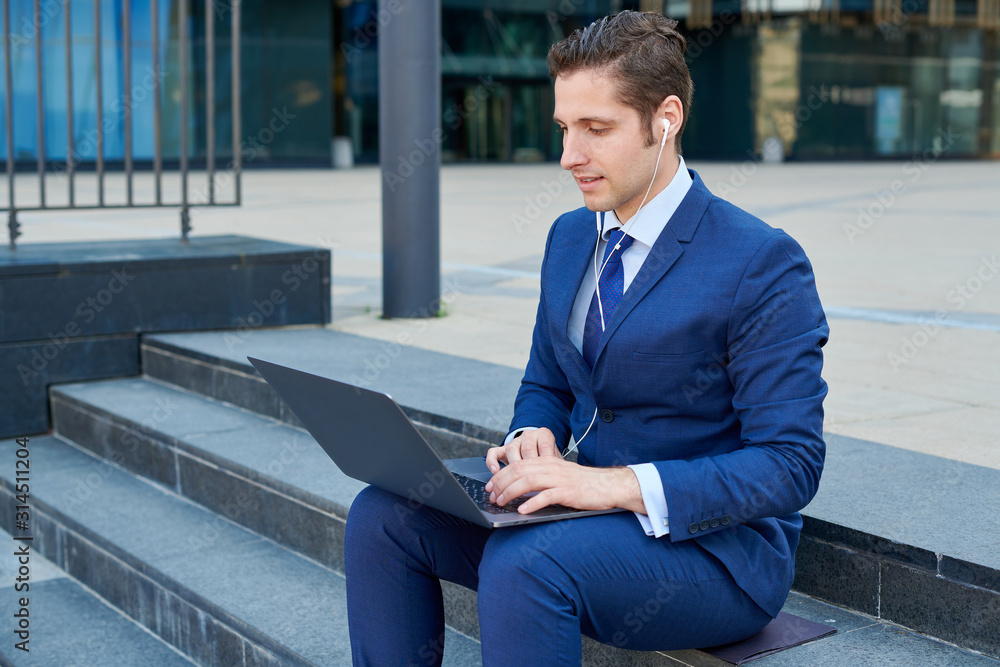 Happy sucessfull young businessman working on modern laptop, using earphones and sitting on stairs outside skyscapers Happy sucessfull young businessman working on modern laptop, using earphones and sitting on stairs outside skyscapers