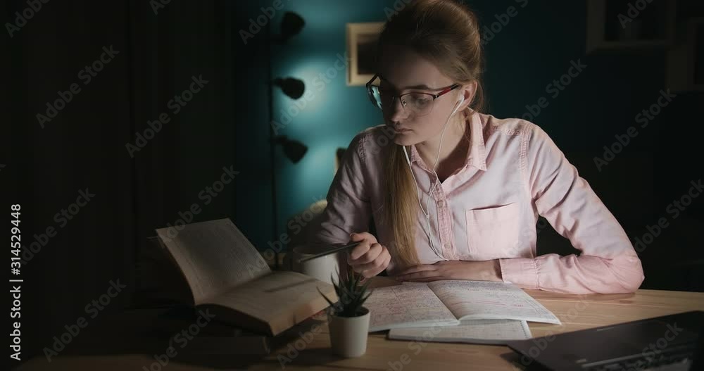 Young attractive lady student in spectacles and earphones studying late evening at home, book warm Young attractive lady student in spectacles and earphones studying late evening at home, book warm