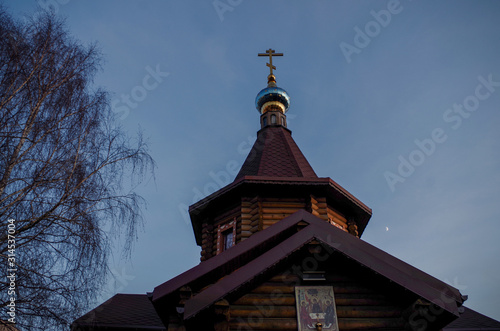Gable roof of wooden Orthodox Church with log walls, six-sided tower, blue dome and gilded Orthodox cross with an icon above entrance on the background of evening blue sky and birch tree on the left