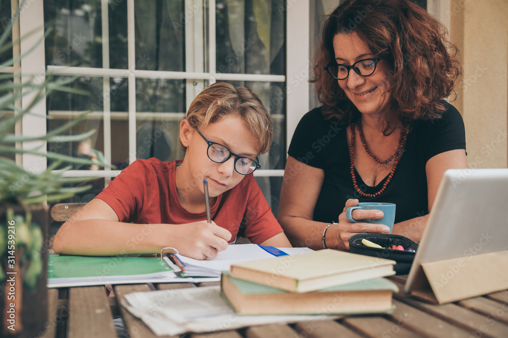 Obraz premium Young student doing homework at home with school books newspaper, digital pad helped by his mother. Mum control, help and teaching his son. Education, family, lifestyle and homeschooling concept.