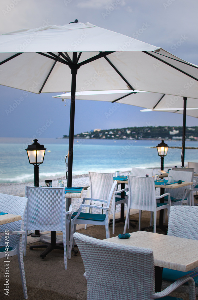 Tables for a romantic meal on the beach with lanterns and chairs. With sunset view. Menton, France