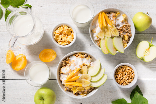 Bowls with granola, fruit, yogurt and two glasses with milk on a white wooden background. Healthy breakfast cereal