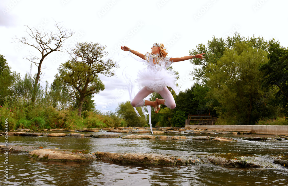 A girl is dressed up as a fairy all in white. She is seen alongside of a flowing river  with large stepping stones. 