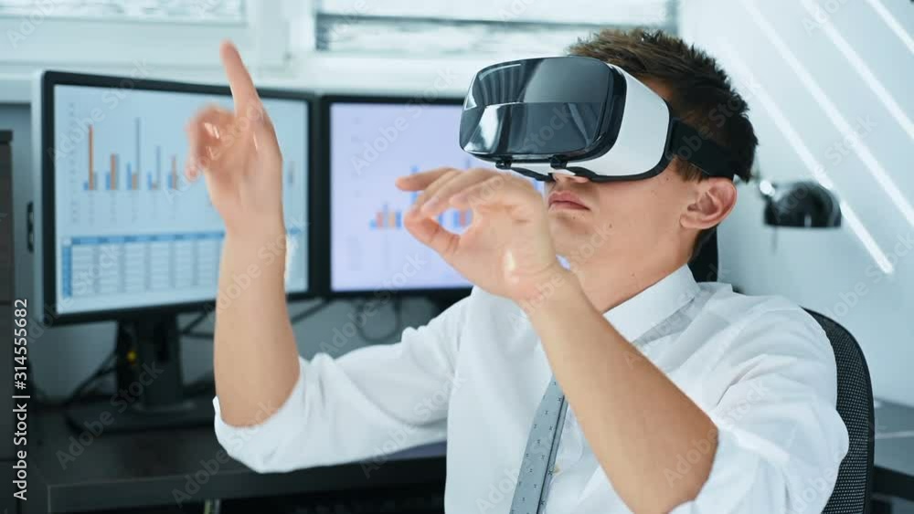 Trader Man With VR Headset Sitting In Office And Looking On Virtual Stock Chart. Business And Communication Concept Trader Man With VR Headset Sitting In Office And Looking On Virtual Stock Chart. Business And Communication Concept