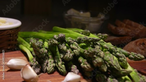 Peeled green asparagus spear falls onto a wooden board next to tha garlic and cheese photo