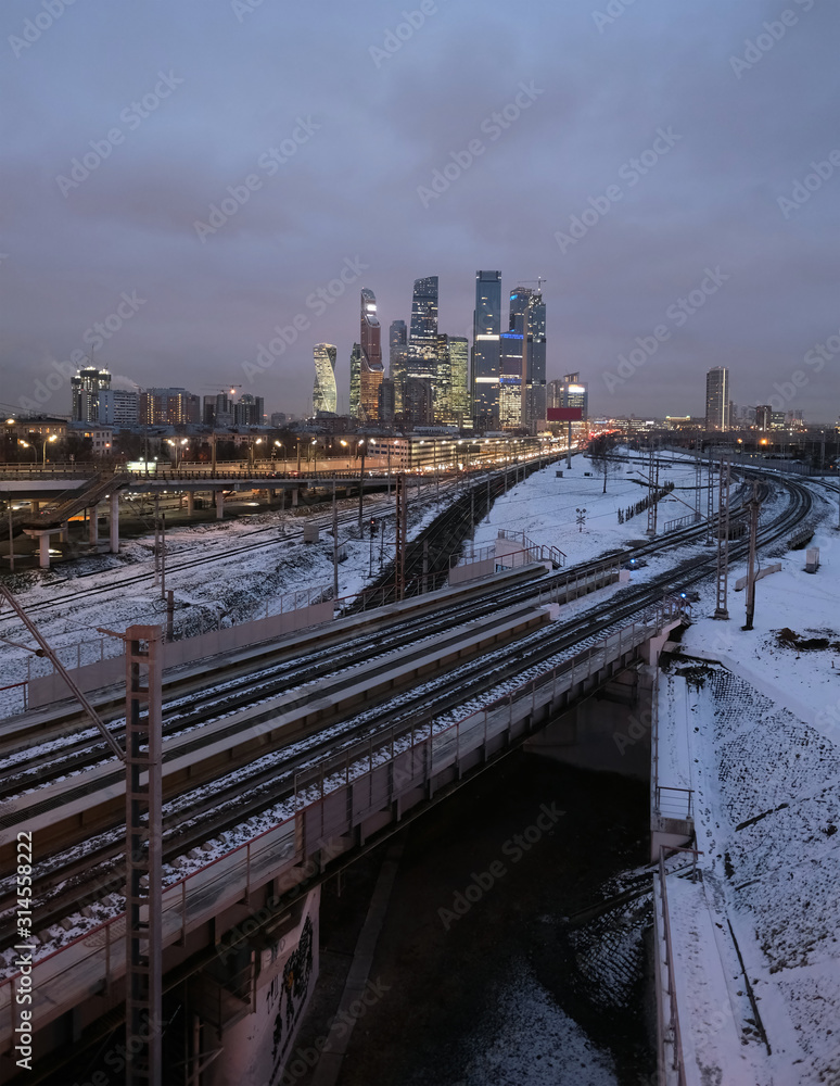 Industrial landscape with many railroad ways going far and skyscrapers with power plant pipes on horizon at night in winter period vertical view Industrial landscape with many railroad ways going far and skyscrapers with power plant pipes on horizon at night in winter period vertical view