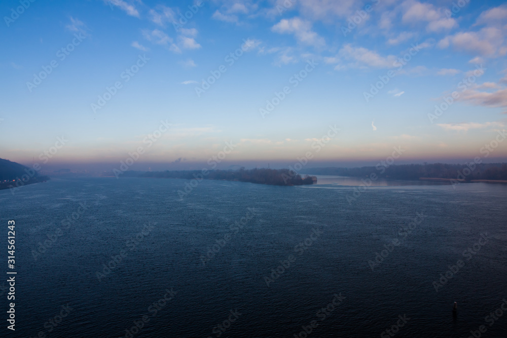 Fototapeta premium Cold early winter morning, light haze and fog rise above the calm water of the river, on the shore there are trees without leaves.Bridge and the silhouette of the buildings in the distance. Kyiv.