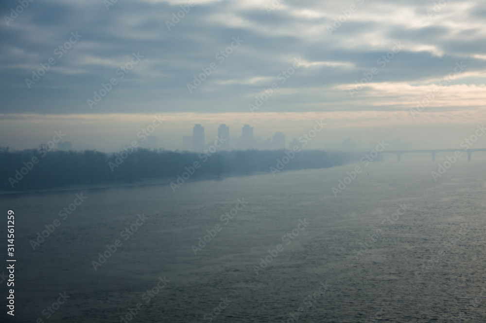 Fototapeta premium Cold early winter morning, light haze and fog rise above the calm water of the river, on the shore there are trees without leaves.Bridge and the silhouette of the buildings in the distance. Kyiv.