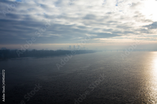 Cold early winter morning, light haze and fog rise above the calm water of the river, on the shore there are trees without leaves.Bridge and the silhouette of the buildings in the distance. Kyiv. 