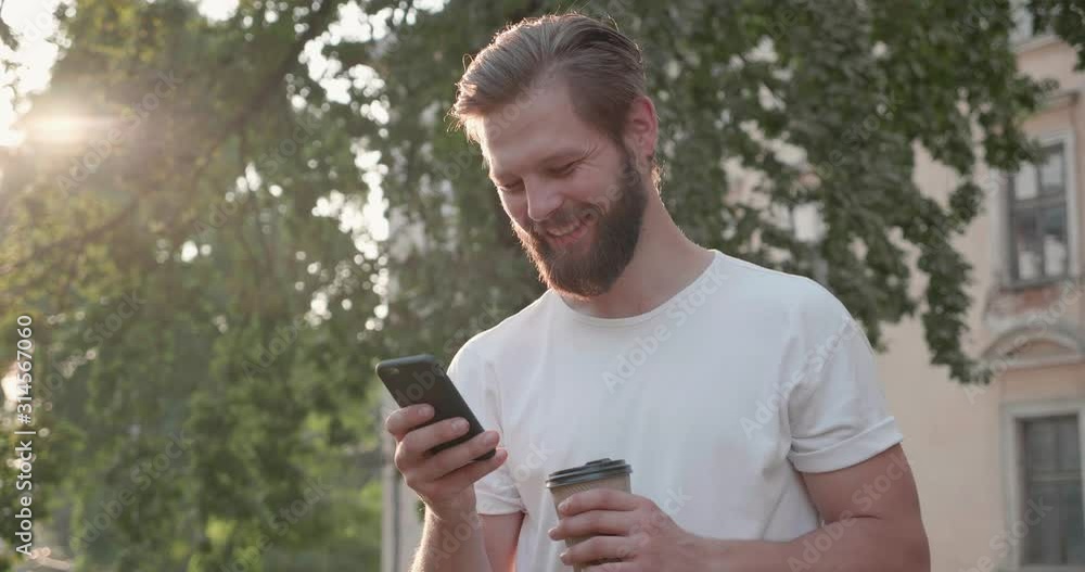 Portrait of Bearded Athletic Man Wearing white T-shirt Standing in the Town with Mobile Phone. Having online Conversation on it. Looking stylish while having well groomed Hairstyle. Sunny day Portrait of Bearded Athletic Man Wearing white T-shirt Standing in the Town with Mobile Phone. Having online Conversation on it. Looking stylish while having well groomed Hairstyle. Sunny day