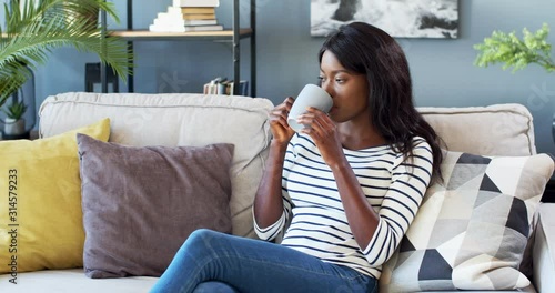 African American young beautiful woman sipping hot tea or coffee while sitting on the couch, looking at the window and dreaming in the cozy living room.