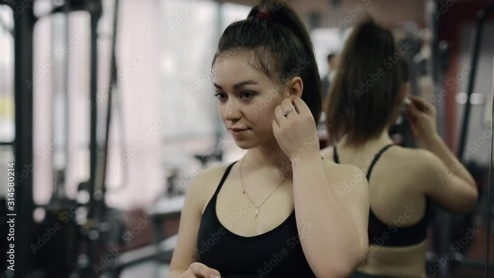 Young woman puts on wireless earbuds with music at the gym ready to workout Young woman puts on wireless earbuds with music at the gym ready to workout