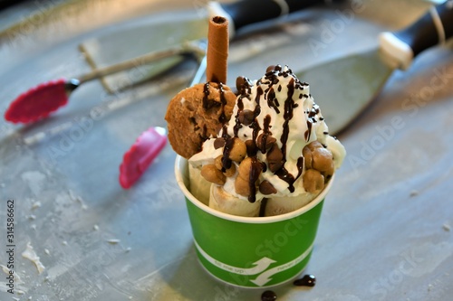 Rolled ice cream dessert in a green cardboard cup with cookie, chocolate sauce, cookie dough and whipped cream with creation tools in the background