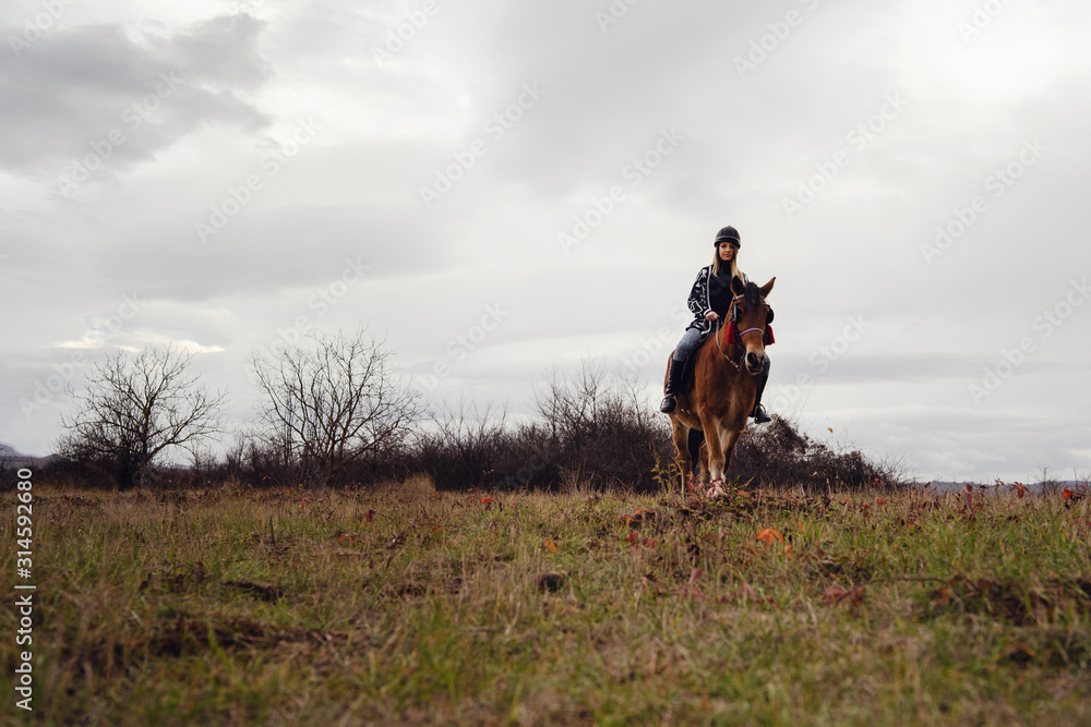 Young caucasian blonde woman female girl on the horse riding in nature wearing helmet in winter or autumn day against a a gray sky in the field