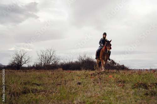 Young caucasian blonde woman female girl on the horse riding in nature wearing helmet in winter or autumn day against a a gray sky in the field © Miljan Živković