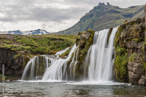 View of three waterfalls. Water falls from small cliffs covered with green grass. Mountains and cloudy sky in the background