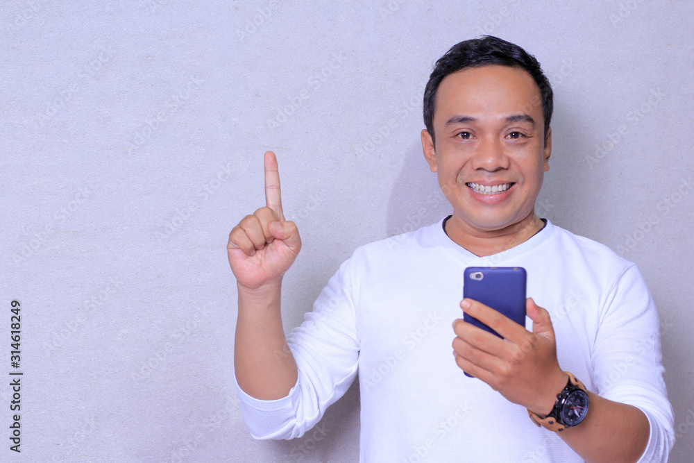 Young Asian man using smartphone standing surprised with an idea or question pointing finger with happy face, number one Young Asian man using smartphone standing surprised with an idea or question pointing finger with happy face, number one