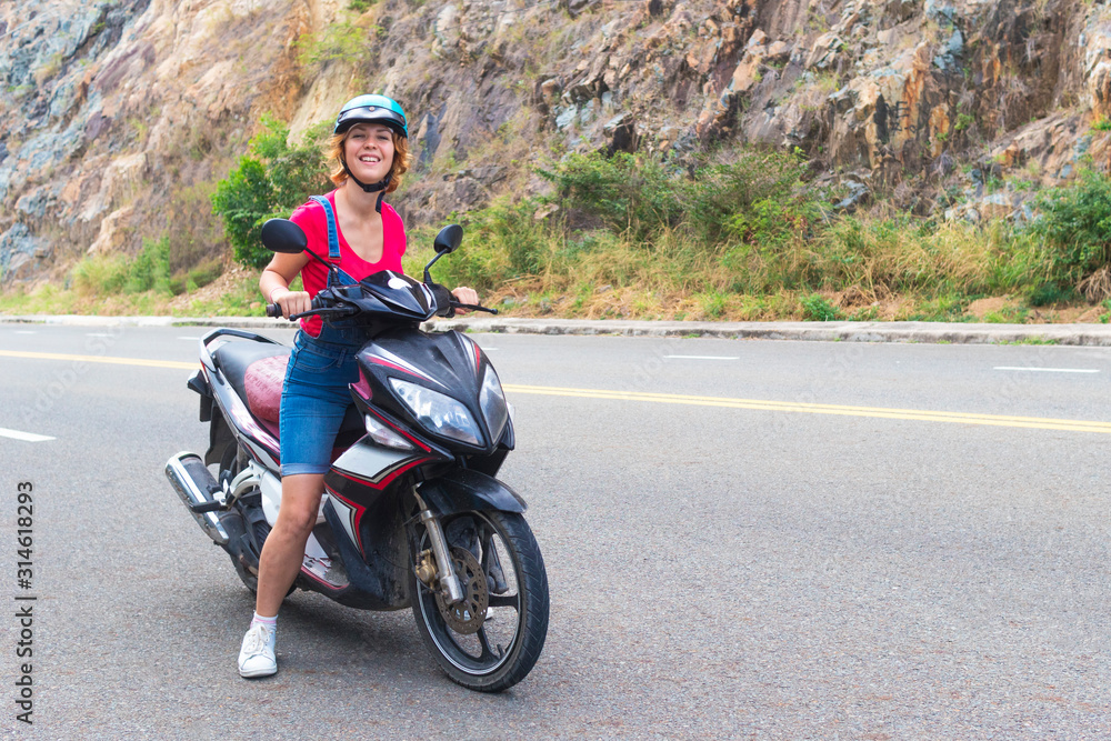 Beautiful happy girl, young woman, biker or motorcyclist  is riding, driving motorcycle, moped or bike, looking at camera, smiling. Female rider  in helmet on the road in mountains in a summer day.