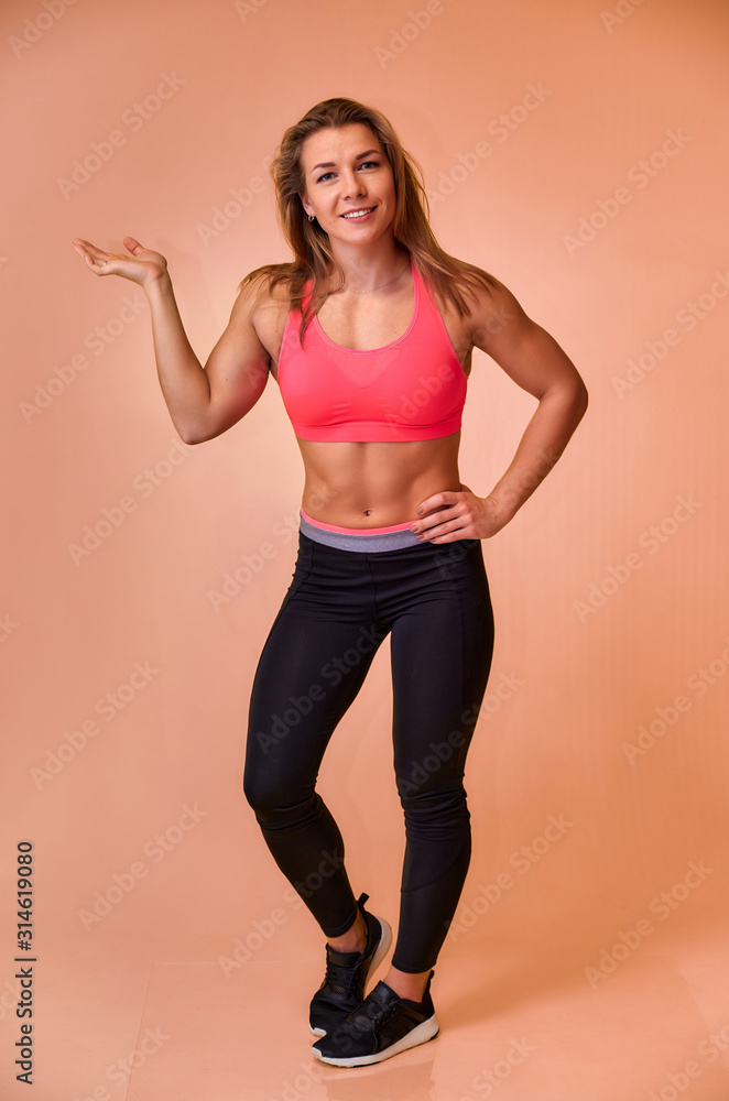 Vertical portrait of a pretty fitness blonde girl with a beautiful smile, excellent teeth and a bodybuilder figure in a tracksuit on a pink background. Cute looks at the camera, standing straight.