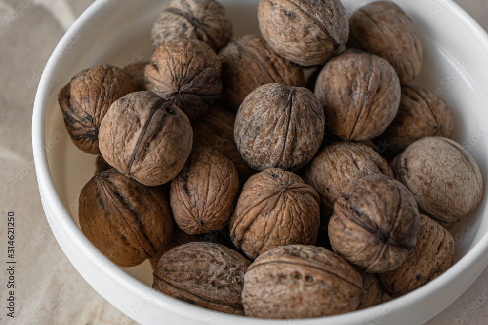 A white plate full of inshell walnuts stands on a brown background from crumpled old kraft paper. Top view flatlay A white plate full of inshell walnuts stands on a brown background from crumpled old kraft paper. Top view flatlay