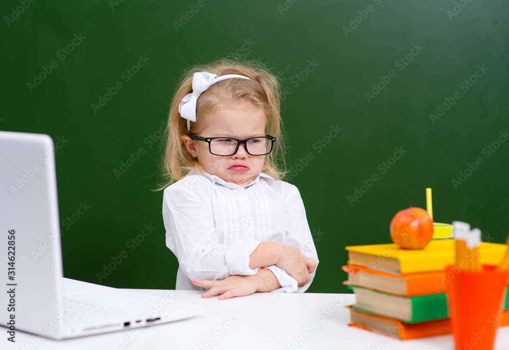 Cute little girl in glasses is working on a laptop at school and showing displeased funny faces Cute little girl in glasses is working on a laptop at school and showing displeased funny faces