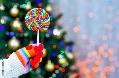 hand in multi-colored glove holds a lollipop against the background of the Christmas tree