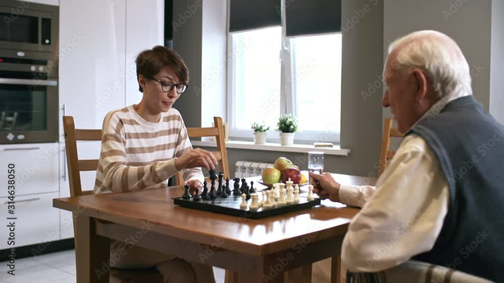 PAN of cheerful young woman in glasses sitting at kitchen table and playing chess with elderly grandfather in wheelchair PAN of cheerful young woman in glasses sitting at kitchen table and playing chess with elderly grandfather in wheelchair