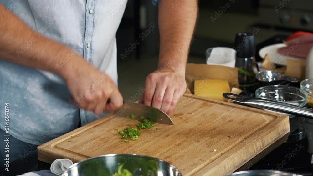 Professional chef stands near kitchen table and expertly slices green seasoning with sharp knife, quick slices, cooks in restaurant, sliced salad, follows recipe, makes breakfast, healthy food Professional chef stands near kitchen table and expertly slices green seasoning with sharp knife, quick slices, cooks in restaurant, sliced salad, follows recipe, makes breakfast, healthy food