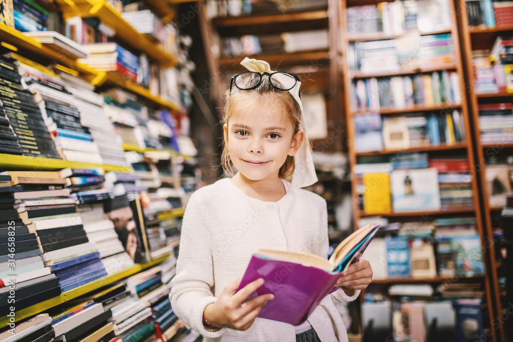 Adorable curious caucasian little girl with eyeglasses standing in bookstore and holding interesting book while looking at camera. All around are books on shelves. Adorable curious caucasian little girl with eyeglasses standing in bookstore and holding interesting book while looking at camera. All around are books on shelves.