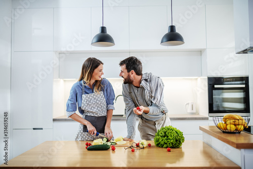 Young charming smiling caucasian woman in apron standing in kitchen and cutting cucumber while talking with her boyfriend. Man holding cherry tomato and talking about healthy lifestyle.