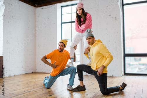 multicultural men in hats holding attractive dancer with crossed arms