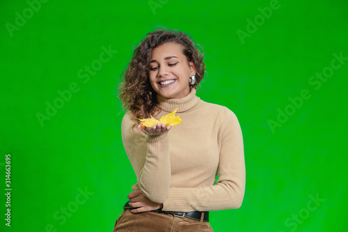 Green background and cheerful afro-american girl posing, bright fallen autumn leaf in her palm.