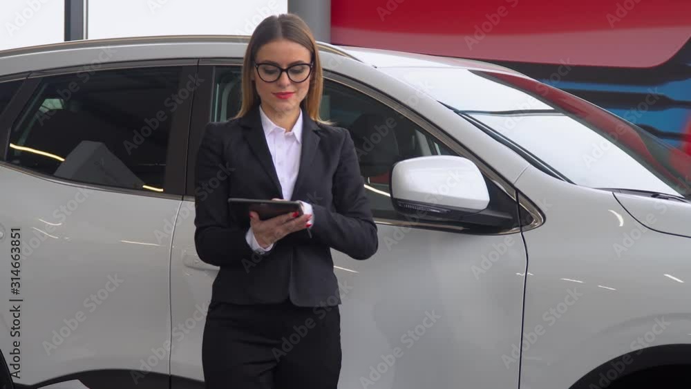 The female seller stands by the car in the car dealership and makes notes on the tablet The female seller stands by the car in the car dealership and makes notes on the tablet