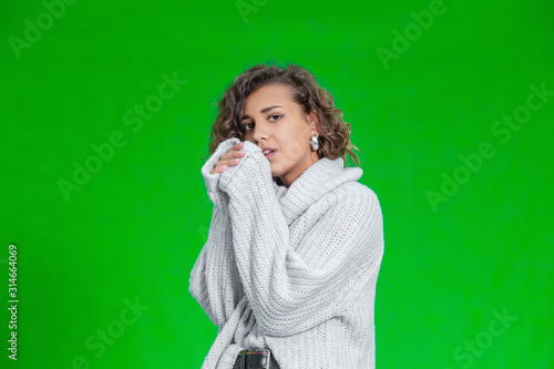Portrait of frozen young female warming hands, standing on green background, weared in warm woolen sweater.