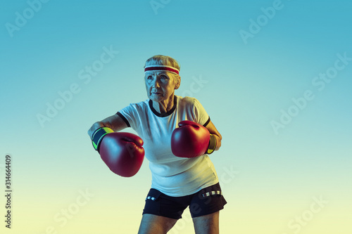 Still kicking. Senior woman in sportwear boxing on gradient background, neon light. Female model in great shape stays active. Concept of sport, activity, movement, wellbeing, confidence. Copyspace.
