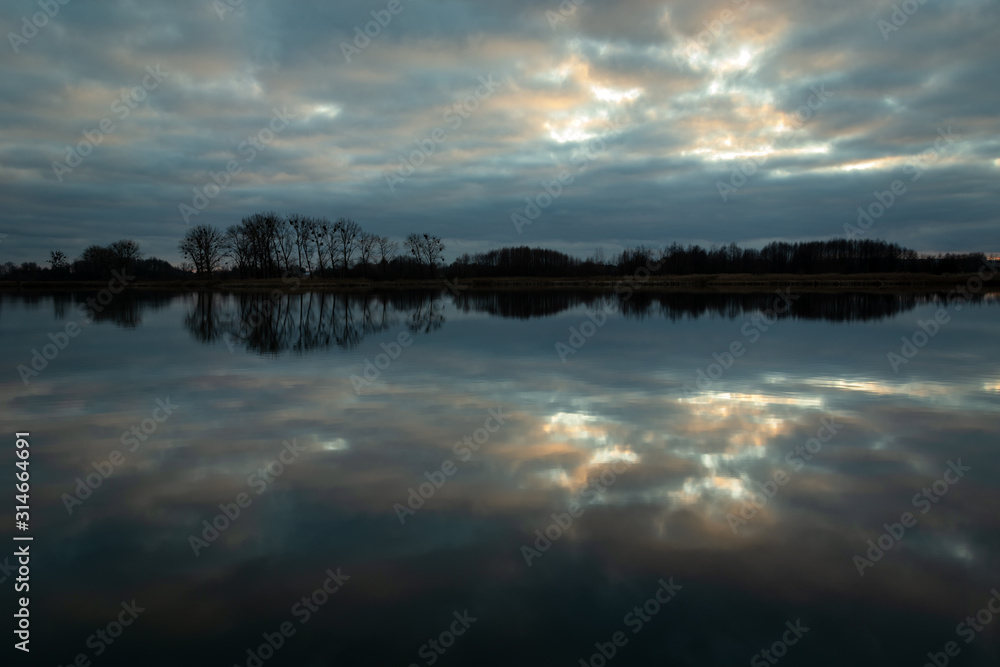 Reflection of clouds in calm water, trees on the horizon, evening view