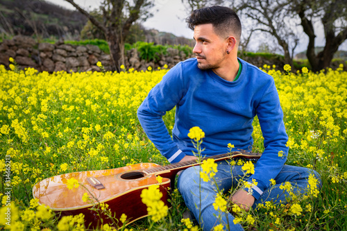 Fotografia Joven músico sentado en un campo de flores con una guitarra acústica apoyada en