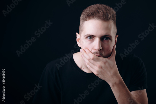 Astonished young man covering his mouth and chin with hand isolated black background