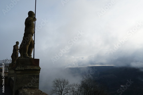 Statues of two Warriors with Speers looking over to the hills surrounding the Hohenzollern castle covered in mist sand clouds in Bisingen Germany. The warriors or guardians are seen in a silhouette. 