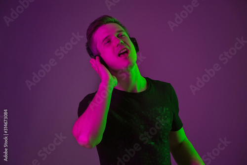 Listen to music, singing, enjoying. Caucasian man's portrait on purple studio background in neon light. Beautiful male model in black shirt. Concept of human emotions, facial expression, sales, ad.