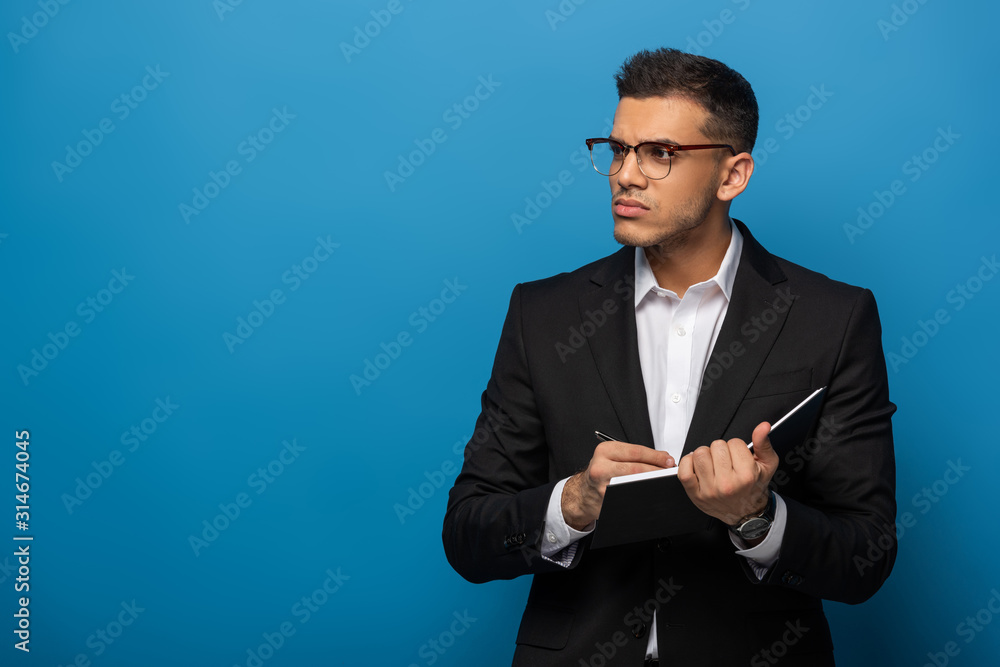 Pensive businessman writing in notebook and looking away on blue background Pensive businessman writing in notebook and looking away on blue background