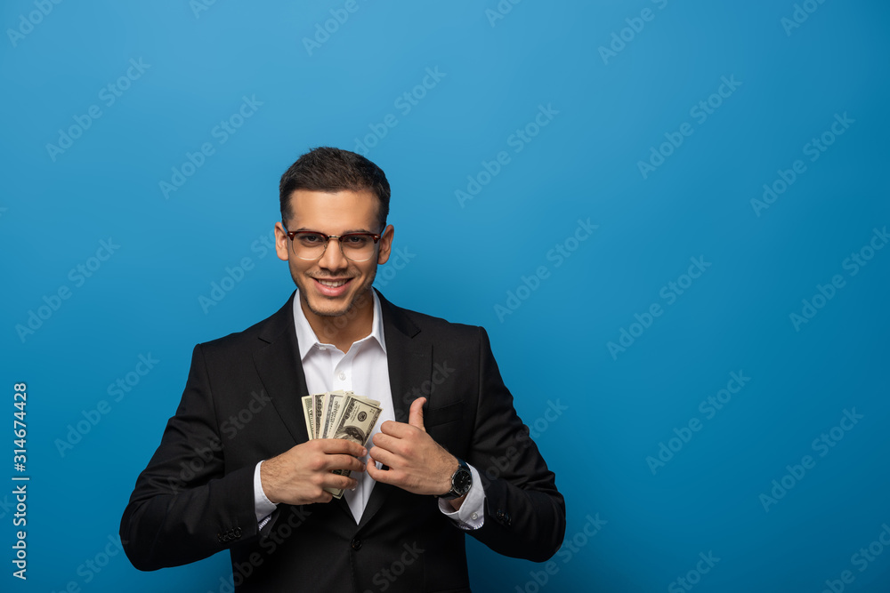 Smiling businessman putting dollar banknotes in jacket pocket on blue background Smiling businessman putting dollar banknotes in jacket pocket on blue background