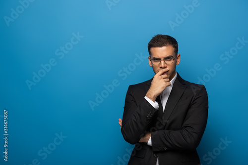 Pensive businessman with hand by chin looking at camera on blue background © LIGHTFIELD STUDIOS